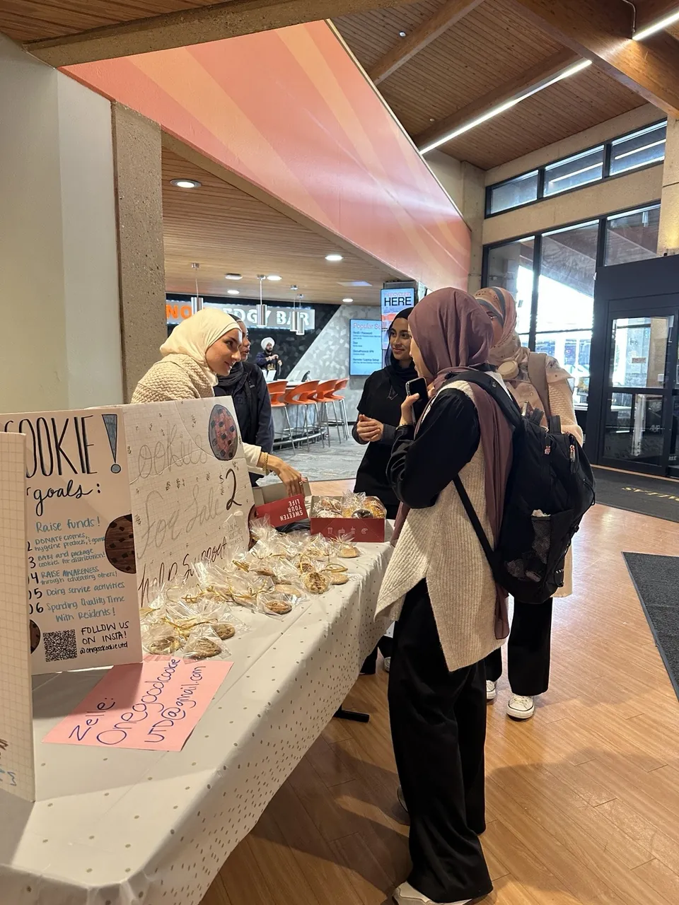 Students stopping by the One Good Cookie bake-sale table on campus, with hand-drawn signs and wrapped cookies.