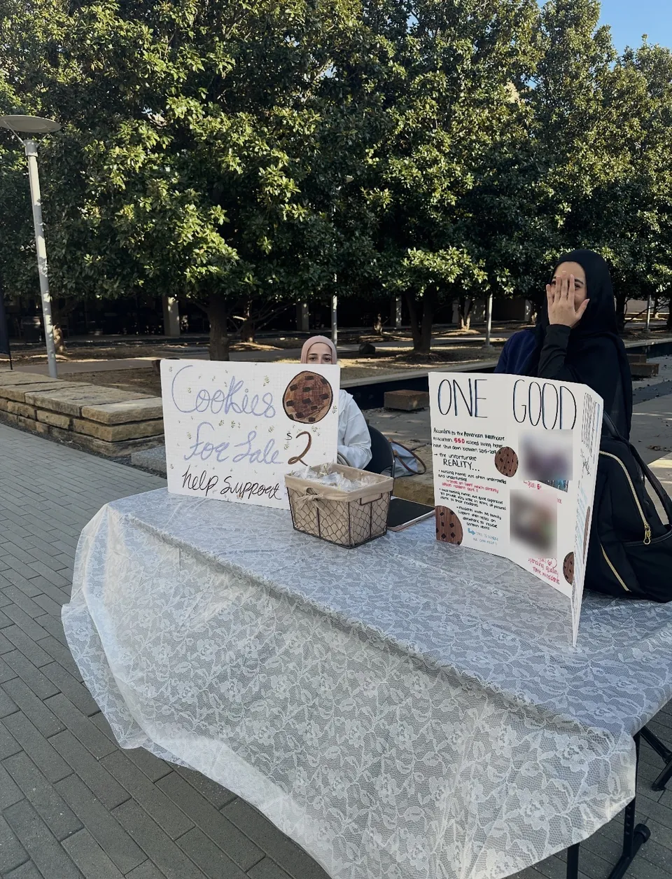Two members sitting behind the One Good Cookie bake-sale table outdoors on campus.