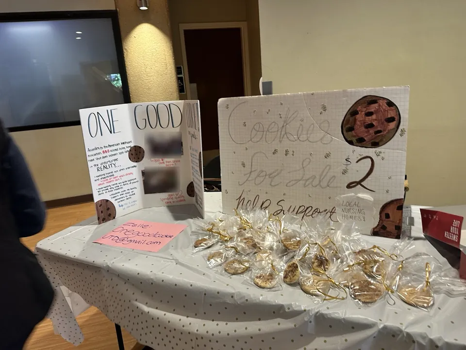 The One Good Cookie bake-sale table with a hand-lettered trifold, a “Cookies for sale” sign, and individually wrapped cookies.